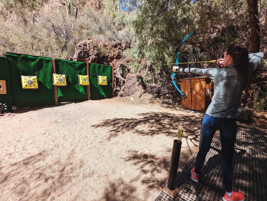 woman with brown hair in a ponytail wearing a dusty green jacket and jeans shooting a blue archery bow towards multiple archery targets a short distance away.