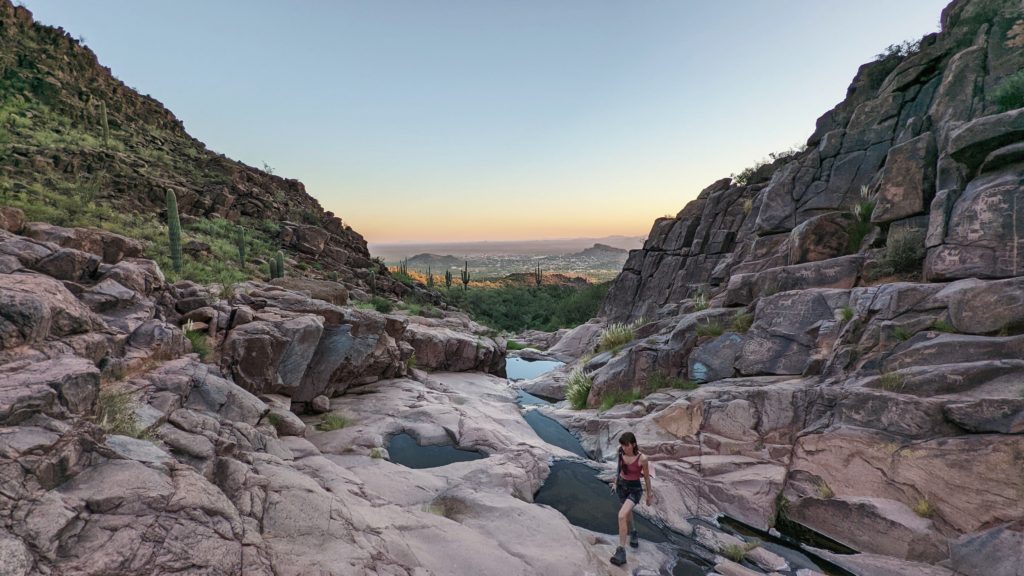 brown haired woman in a pink tank top and grey shorts walking across a rocky stream near the petroglyphs on the Hieroglyphic Trail