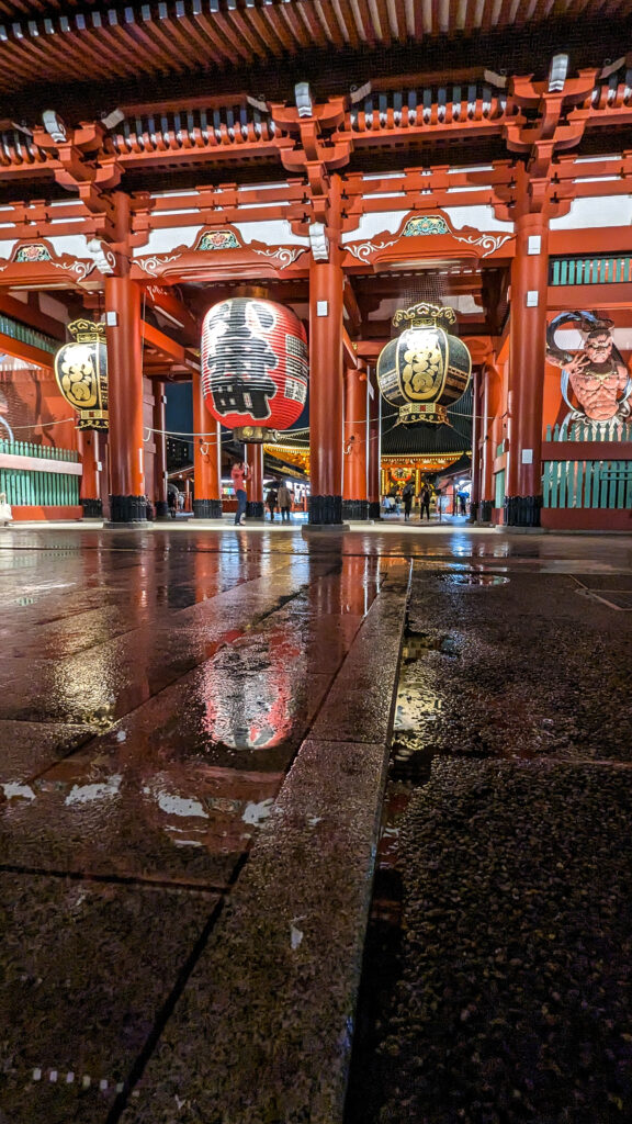 Hozomon gate at Sensoji temple in Japan. Red orange colored ornate gate with large hanging lanterns. It has just rained so the ground is reflecting the lanterns. It's nighttime