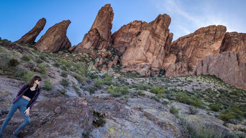 brown haired woman in black sunglass, a black sport bra, purple jacket and grey yoga pants standing in front of a jagged rocky mountain on the treasure loop trail hike.