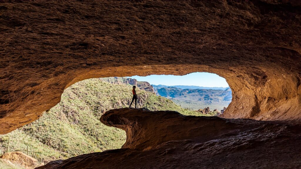 Gabi, The Fringe Explorer, standing on the natural stone wave at the entrance of the wave cave. Beyond the cave is desert landscape and mountains