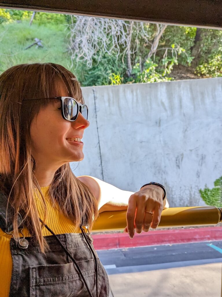 Gabi, The Fringe Explorer, wearing a yellow top and black overalls while sitting on a backlog tour bus