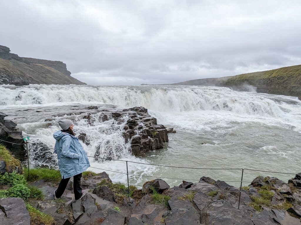 Gabi, The Fringe Explorer, standing in a blue rain jacket with an off white pom pom beanie looking at a large waterfall.