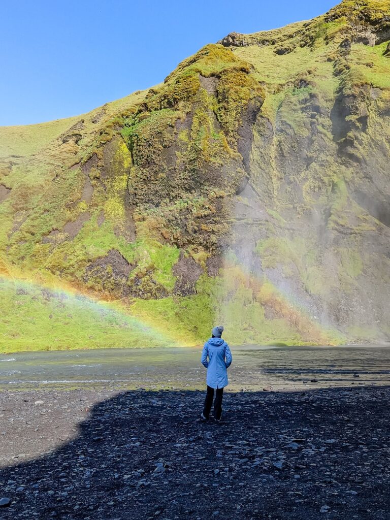 Gabi, The Fringe Explorer, wearing a blue rain coat and beige pom pom beanie while standing next to a stream and double rainbow