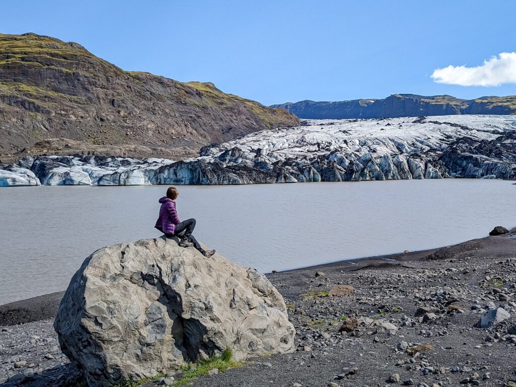 Gabi, The Fringe Explorer, sitting on a rock in front of a glacier. It's a sunny clear day