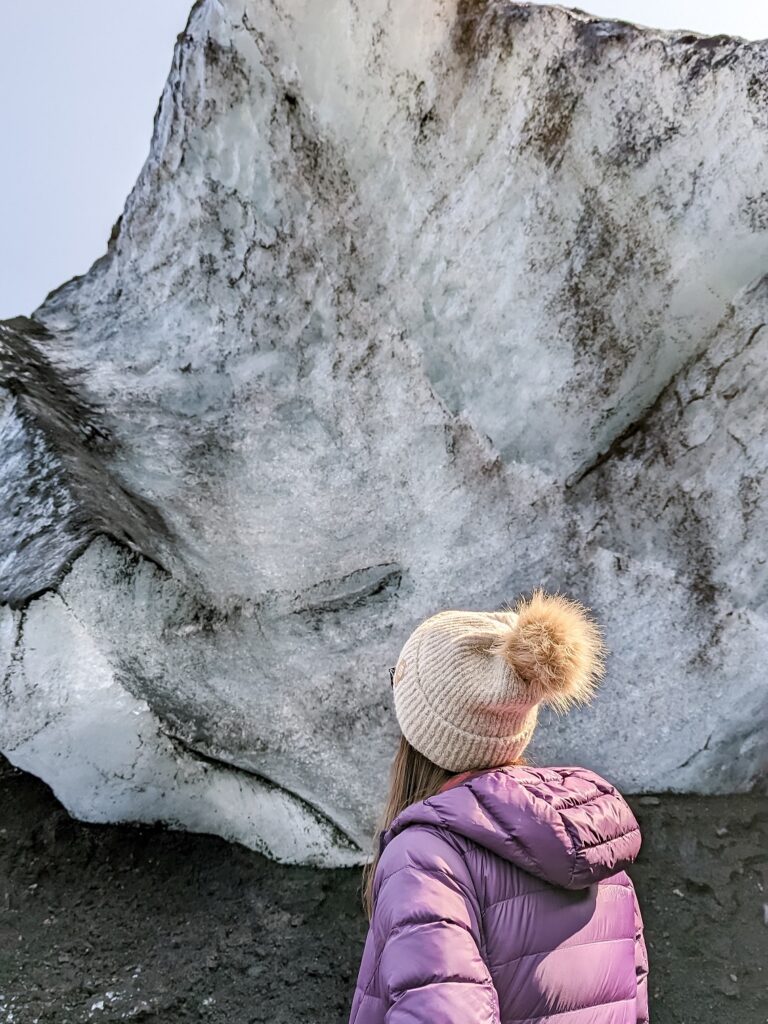 Gabi, The Fringe Explorer, standing next to the wall of a glacier while wearing a purple puffer jacket and beige pom pom benie