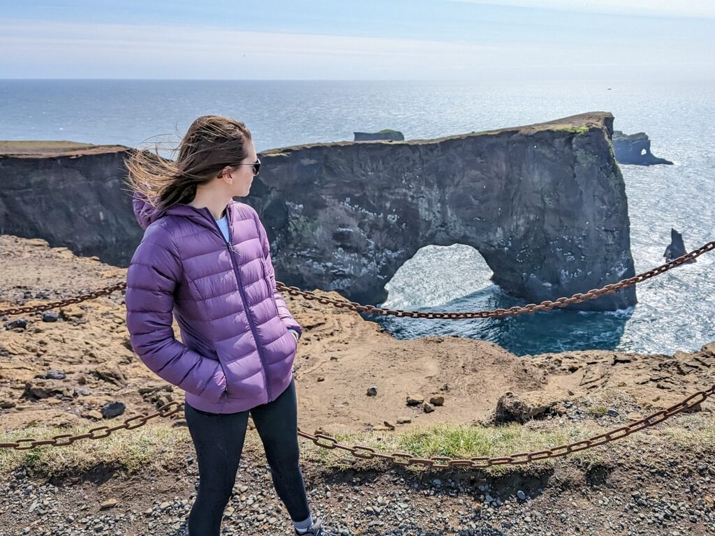 Gabi, The Fringe Explorer, standing in front of a natural stone bridge in the ocean while wearing a purple puffer jacket.