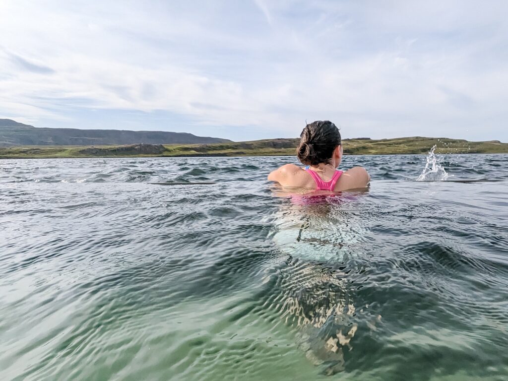 Gabi, The Fringe Explorer, wearing a hot pink bikini top in a hot spring bath, facing a lake