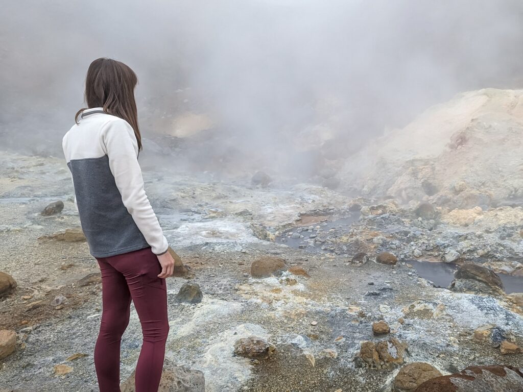 Gabi, The Fringe Explorer, standing in a streamy geothermal field while wearing a grey and white block color pullover and maroon leggings