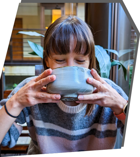 Gabi, a brown haired woman with bangs drinking from a large tea cup partially covering her face.