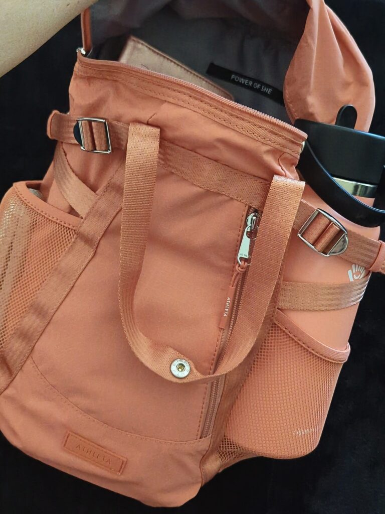 Coral pink Athleta backpack with two water bottle pockets on the side. The bag is unzipped and the right water bottle pocket has a matching coral pink 32ox hydro flask water bottle. 