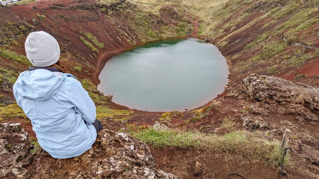 Gabi, The Fringe Explorer, sitting above a blue volcanic crater lake while wearing a light grey beanie and a light blue rain jacket.