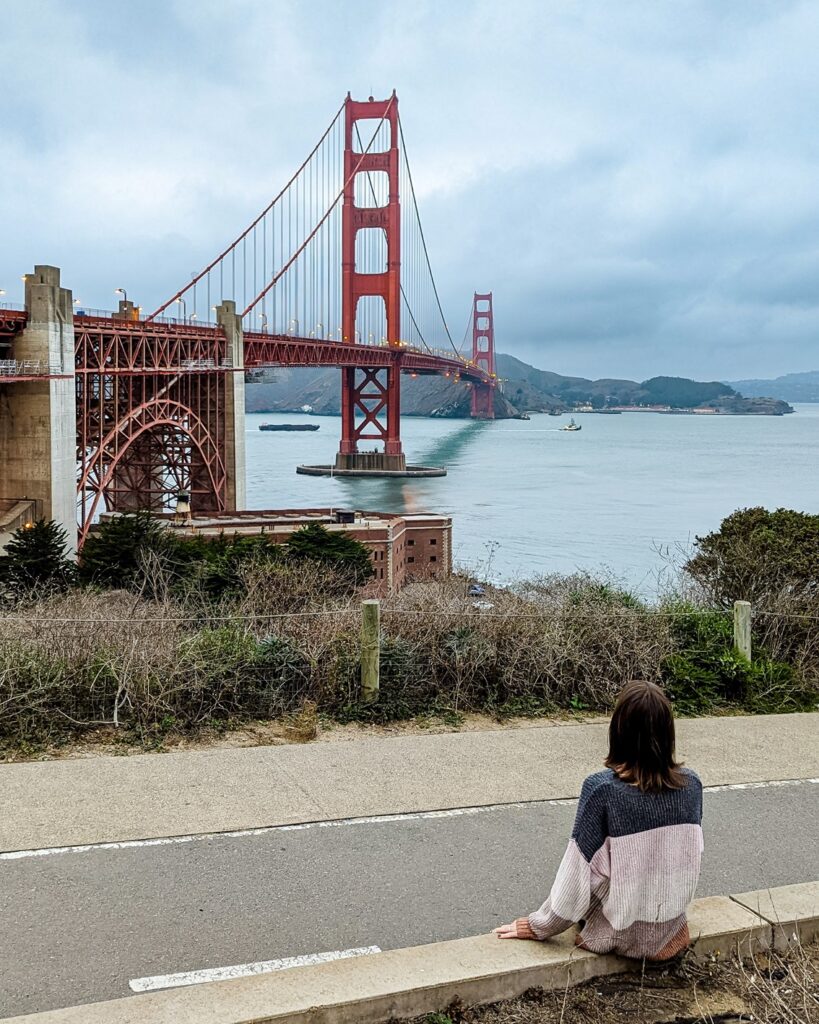 A brown haired girl in a color block sweater is sitting on a ledge looking towards the Golden Gate Bridge in San Francisco