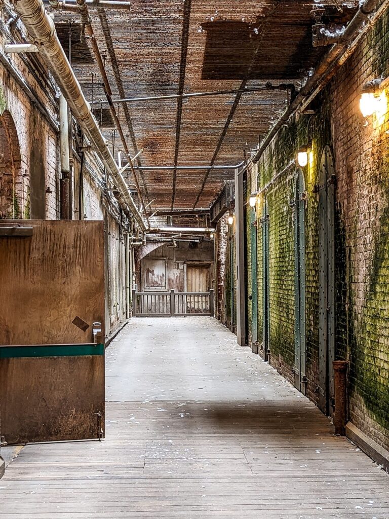 Looking down an empty hall of Alcatraz, the walls are brick, there is a wood door open a third of the way down the hall