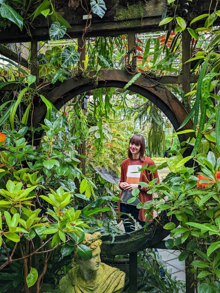 Brown haired woman standing behind a decorative metal-work wall that is covered in plants and vines