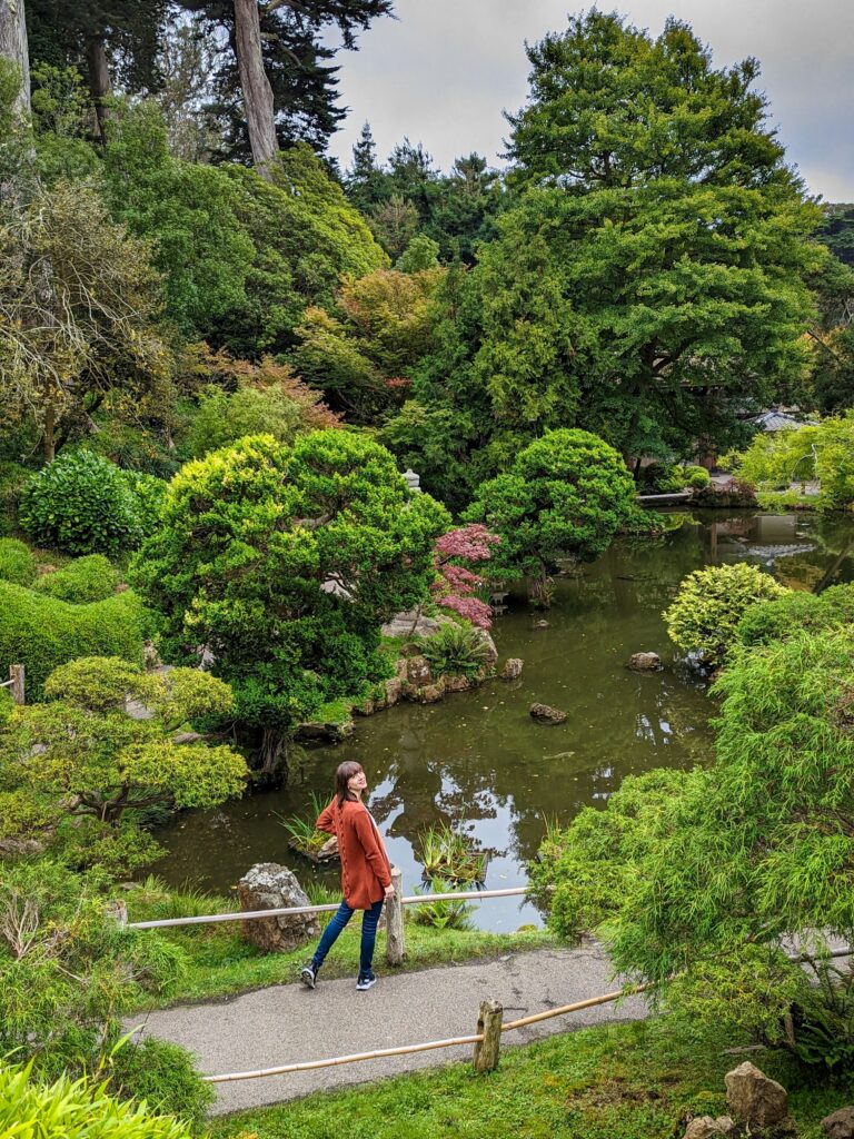 Brown haired girl in a burnt orange sweater looking back towards the camera while standing on a pathway in a Japanese garden never to a pond