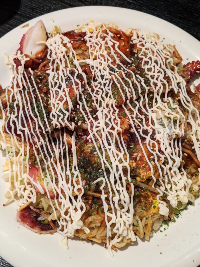 White plate with okonomiyaki - yakisoba noodles topped with cabbage, octopus, thin batter savory pancake, okonomiyaki sauce, Japanese mayonnaise, and seaweed flakes.