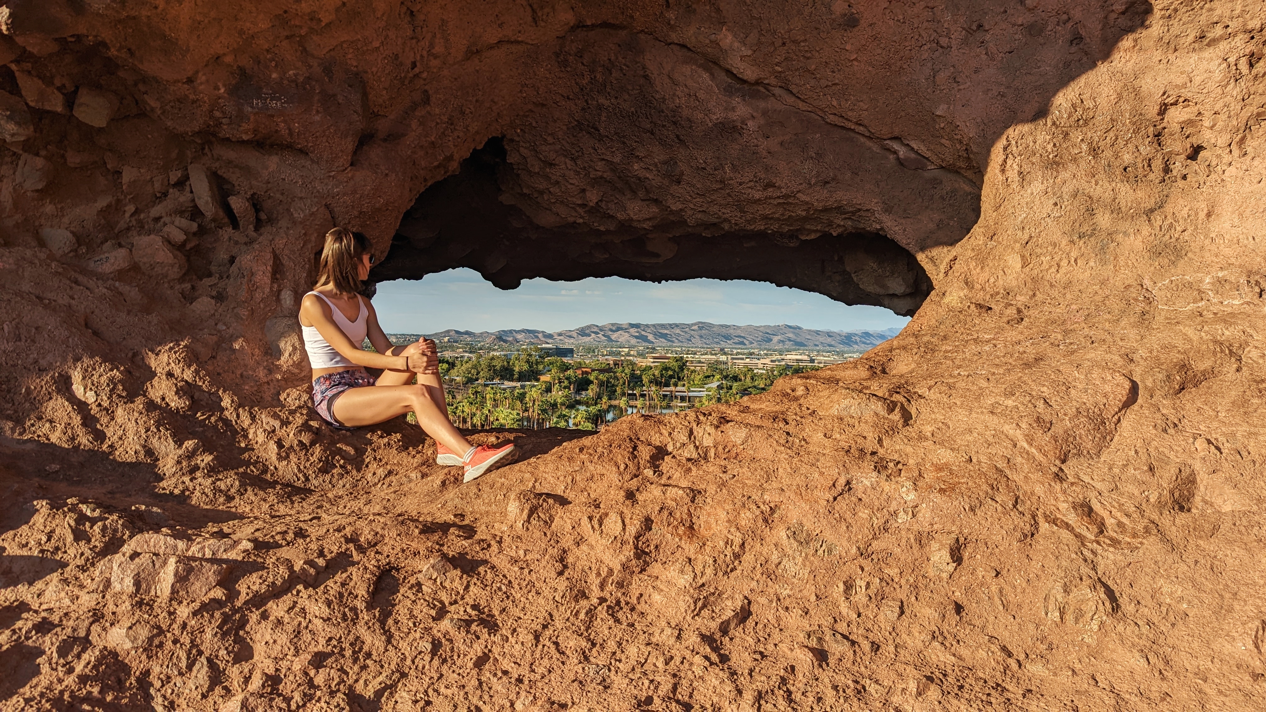 Brown haired woman wearing sunglasses, white tank top, multicolor purple shorts and coral colored shoes looking through a hole in a mountain at a park and city