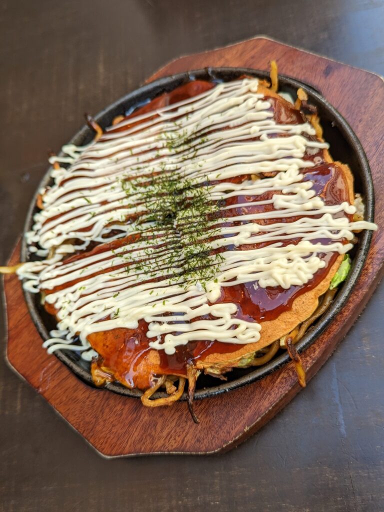 Wood tray with metal dish - inside the metal dish is okonomiyaki. Yakisoba noodles topped with cabbage, thin batter savory pancake, okonomiyaki sauce, Japanese mayonnaise, and seaweed flakes.