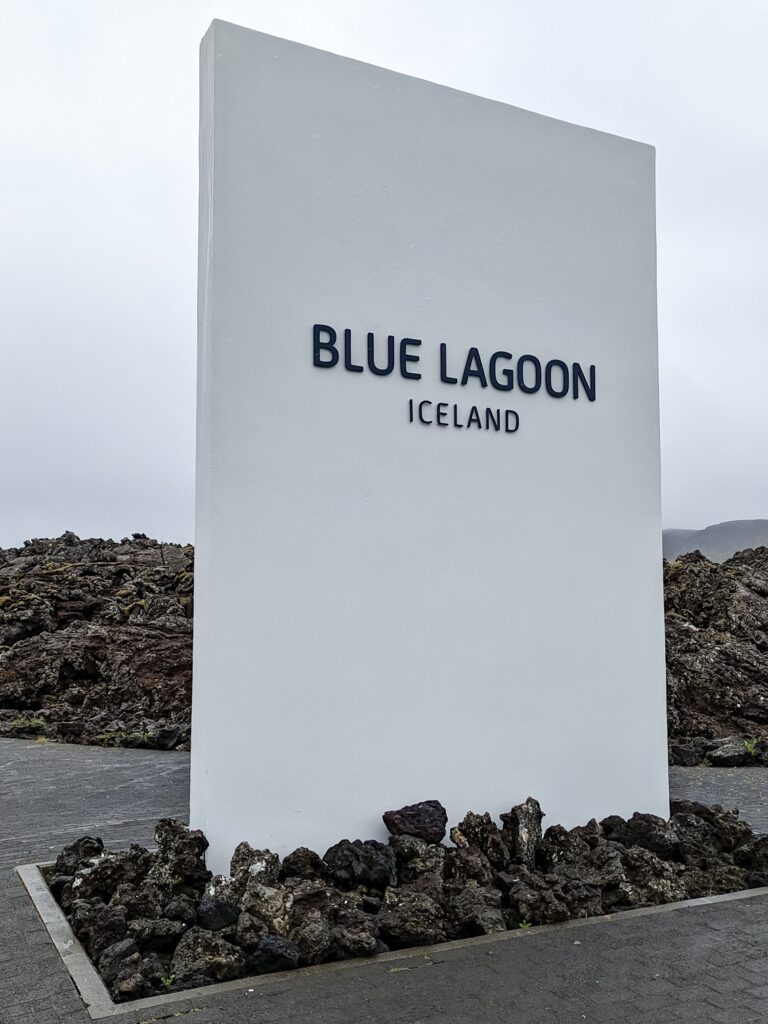 Large white concrete sign standing in the ground surrounded by lava rocks. The sign says "BLUE LAGOON"