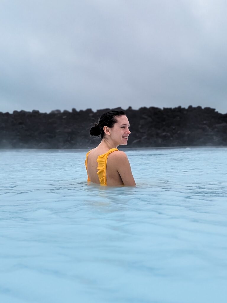 Woman with brown hair in a low bun and yellow swimsuit standing in milky blue waters at the Blue Lagoon.