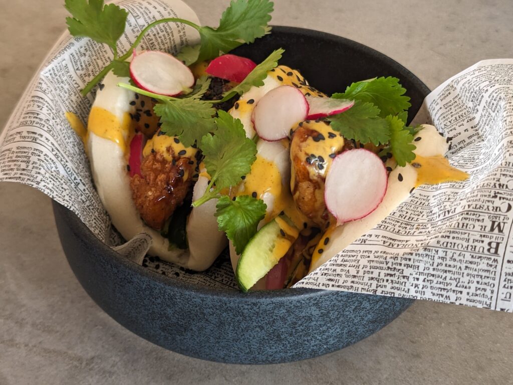 Two bao bun tacos sitting in a shallow grey cylinder-style bowl. The bao have chicken, radishes and greens