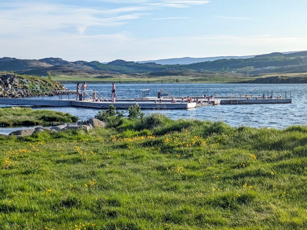 Looking out across a patch of green grass with small yellow flowers at two hot spring baths "floating" on a lake. Many people are soaking int he baths and walking on the wood walkways around the baths.