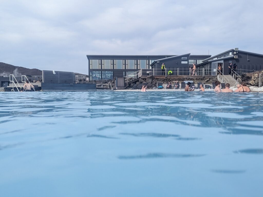 Looking towards a grey two story building with lots of windows across milky blue hot spring waters. people are at the edge of the water near the building