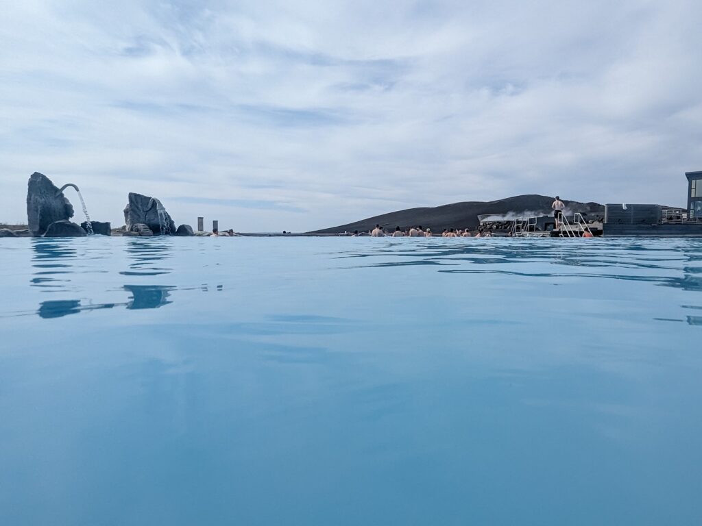 Looking towards a mountain in the distance across milky blue hot spring waters. people are at the edge of the water 