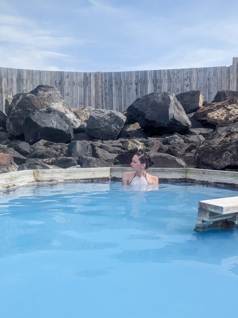 Woman with brown hair pulled back and a crocheted bikini top sits in milk blue hot spring water with large volcanic rocks in the background