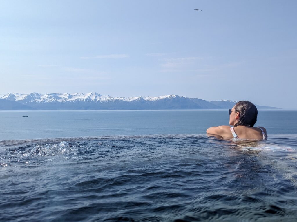 A woman with brown hair pulled back and a pastel stripe bikini top floats againstan infinity edge hot spring pool looking towards snow capped mountains across an ocean in the background