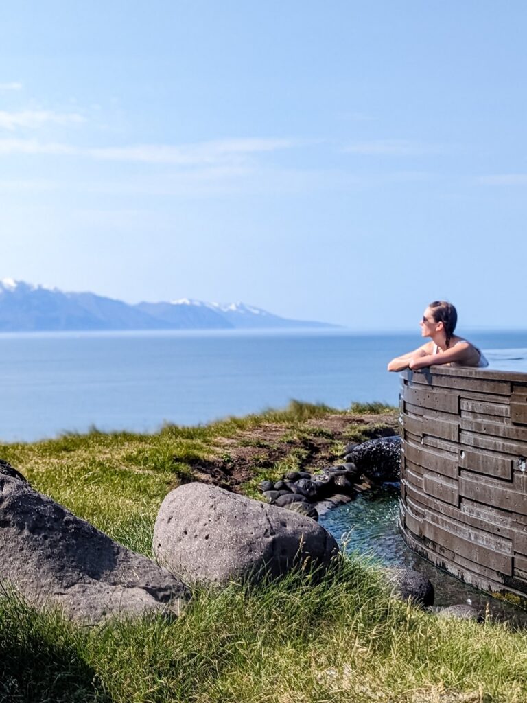 A woman with brown hair pulled back and a vertical pastel stripe bikini top sits in an infinity edge hot spring pool arms crossed looking towards snow capped mountains across an ocean in the background