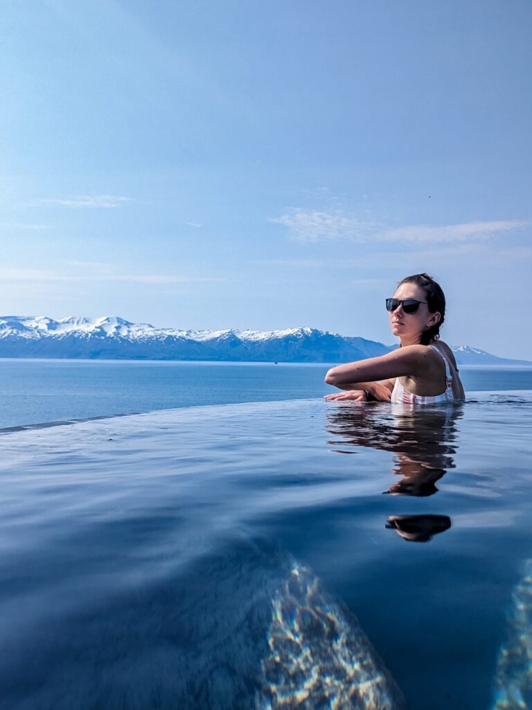 A woman with brown hair pulled back and a vertical pastel stripe bikini top sits in an infinity edge hot spring pool arms crossed looking towards the camera. There are snow capped mountains across an ocean in the background