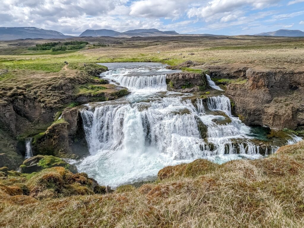 multi-tiered waterfall with turquois waters surrounded by mossy baren landscape with some mountains far off in the background