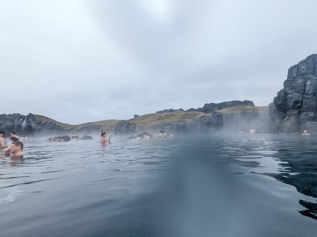 geothermal hot spring surrounded by grey lava rock and moss. Many people are in the background wading in the hot spring.