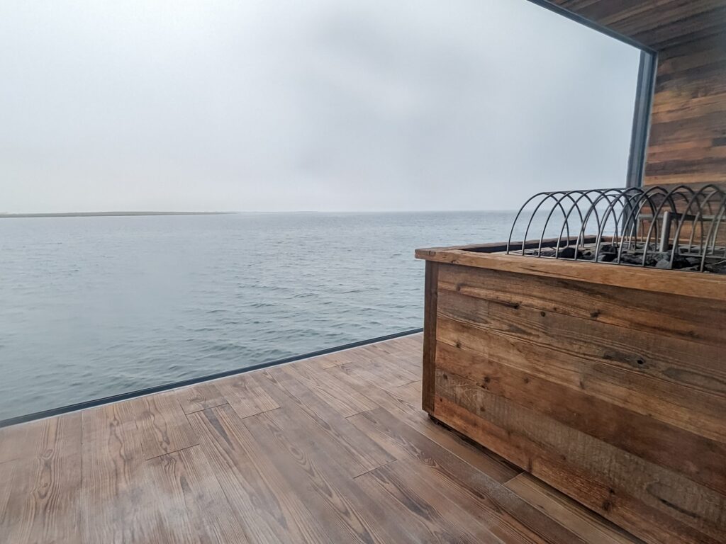 looking out a large glass window at the ocean while sitting inside a brown wood sauna. The sky outside is cloudy and grey