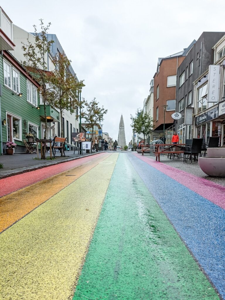 Road painted with a rainbow lines with shops and restaurants, int he distance there is a church spire.