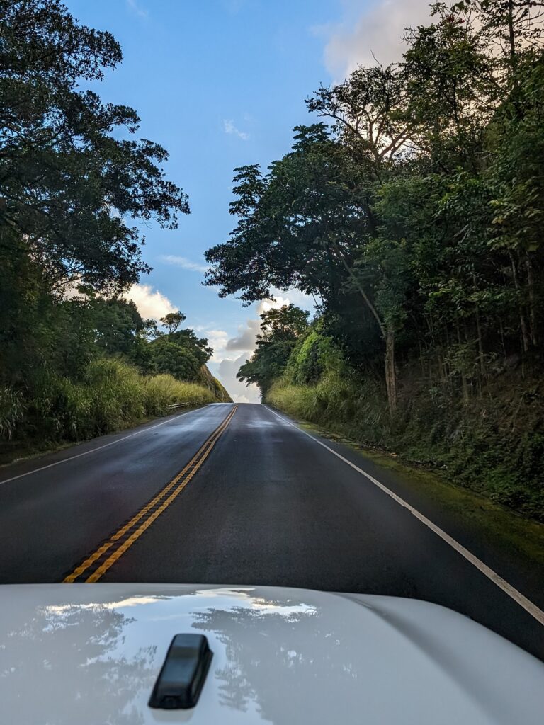 Two lane highway on the "Road to Hana" lined with tropical trees and plants. Clouds on the horizon in the distance.