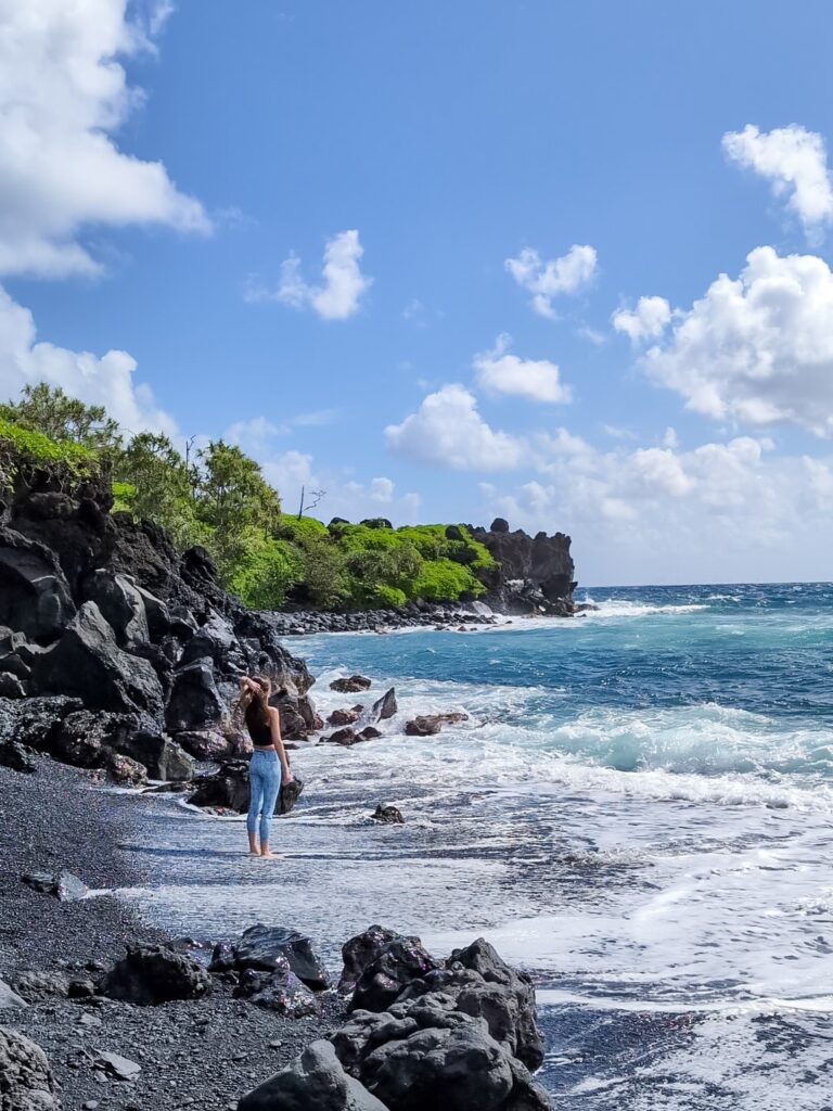 brown-haired woman in sunglasses standing on a black sand beach holding her hair and looking out at the ocean. She is wearing a black cropped tank top and teal leggings with white pineapple print. In the background there is a black rocky area with topical plants growing on top