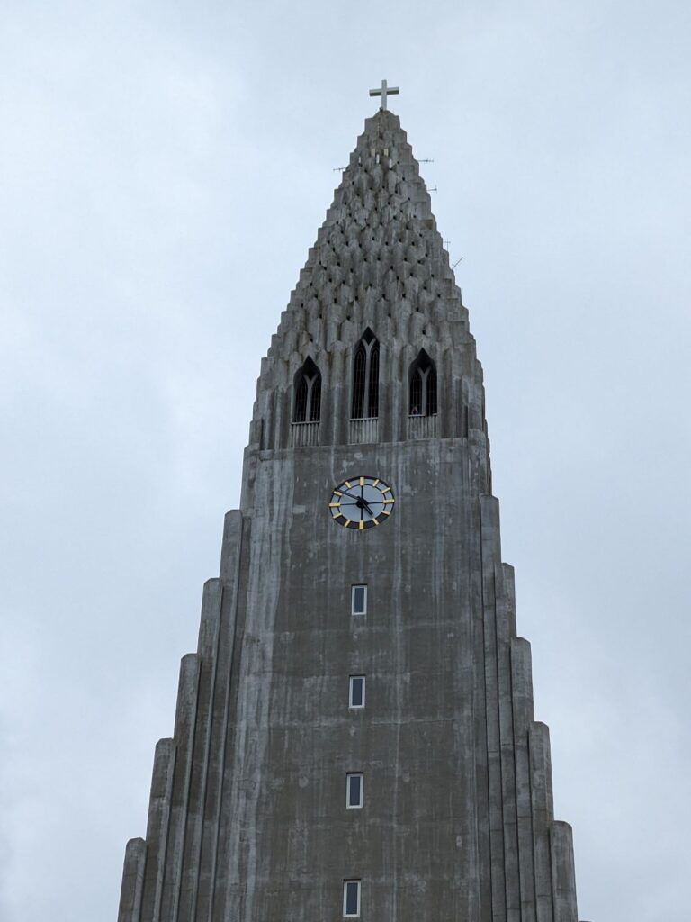 Top of a church spire. Church made made of grey stone with a staircase like architecture.  Near the top there is a clock and then windows above the clock. There is a cross on the very top.