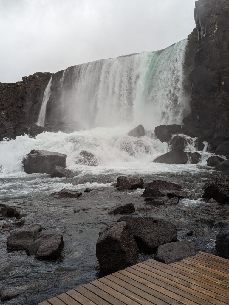 Blue waterfall spilling over a cliff into a rocky river. There is a brown wood deck in the foreground where you can stand.