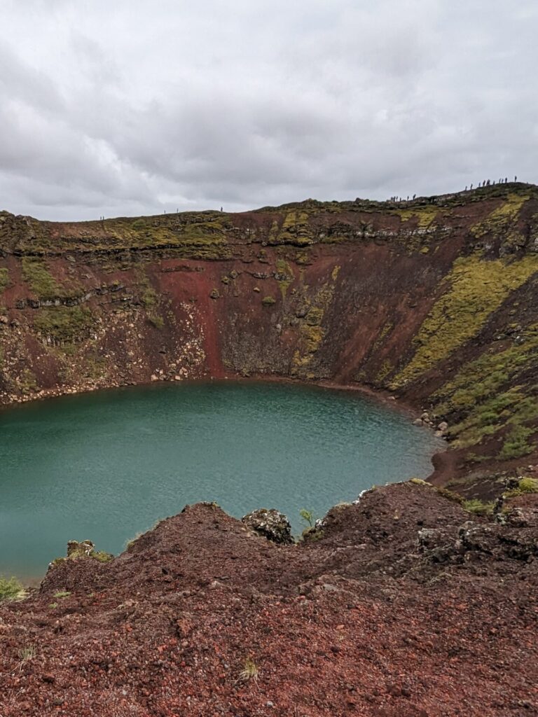 Turquoise lake at the bottom of a red volcanic crater. There are people walking the perimeter int he distance. The sky is cloudy and grey