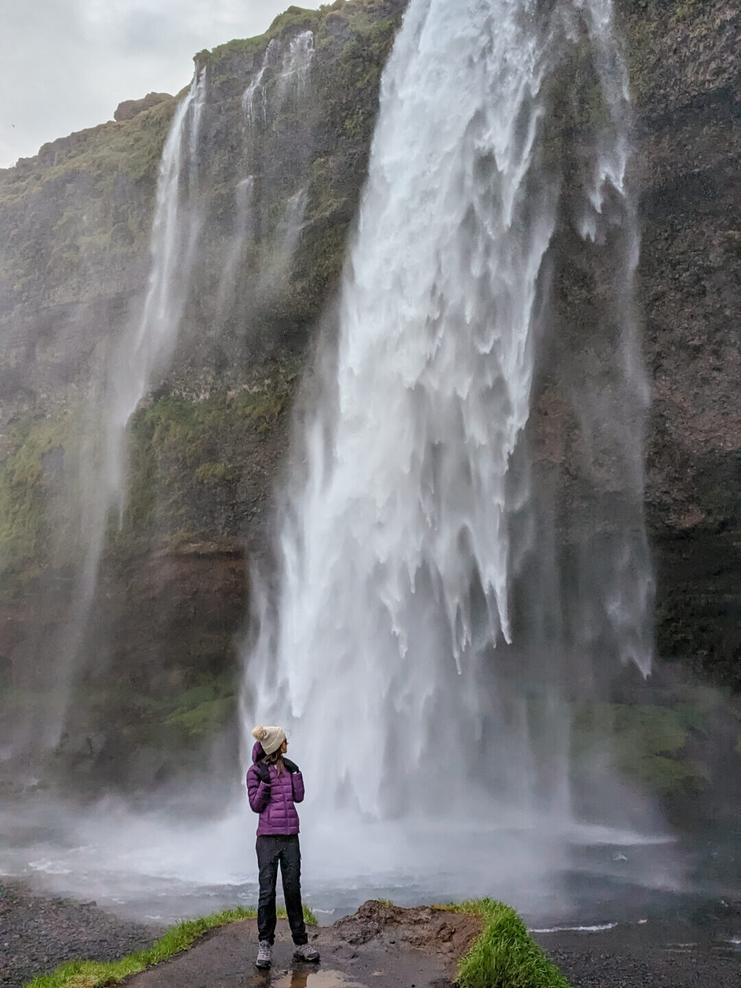 Gabi, a woman wearing a purple puffer jacket, grey hiking pants, hiking boots, and a cream pom pom beanie is standing in front of a waterfall falling over a mossy cliff