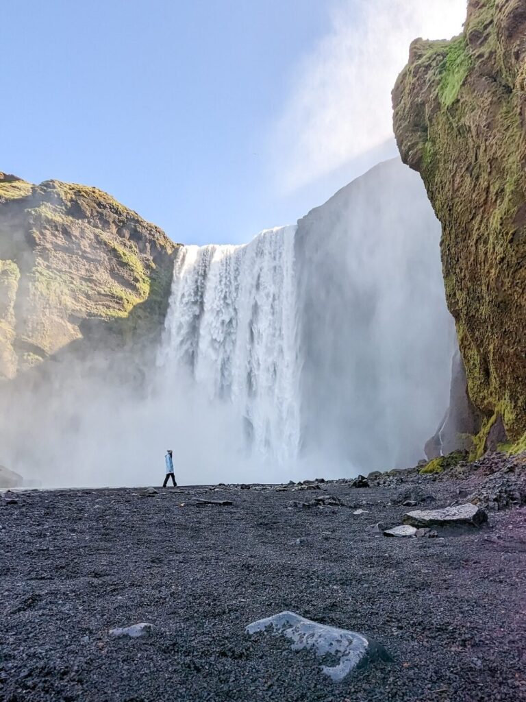 Gabi a woman wearing a blue rain jacket, grey pants, and a pom pom beanie is walking in the distance in front of a waterfall