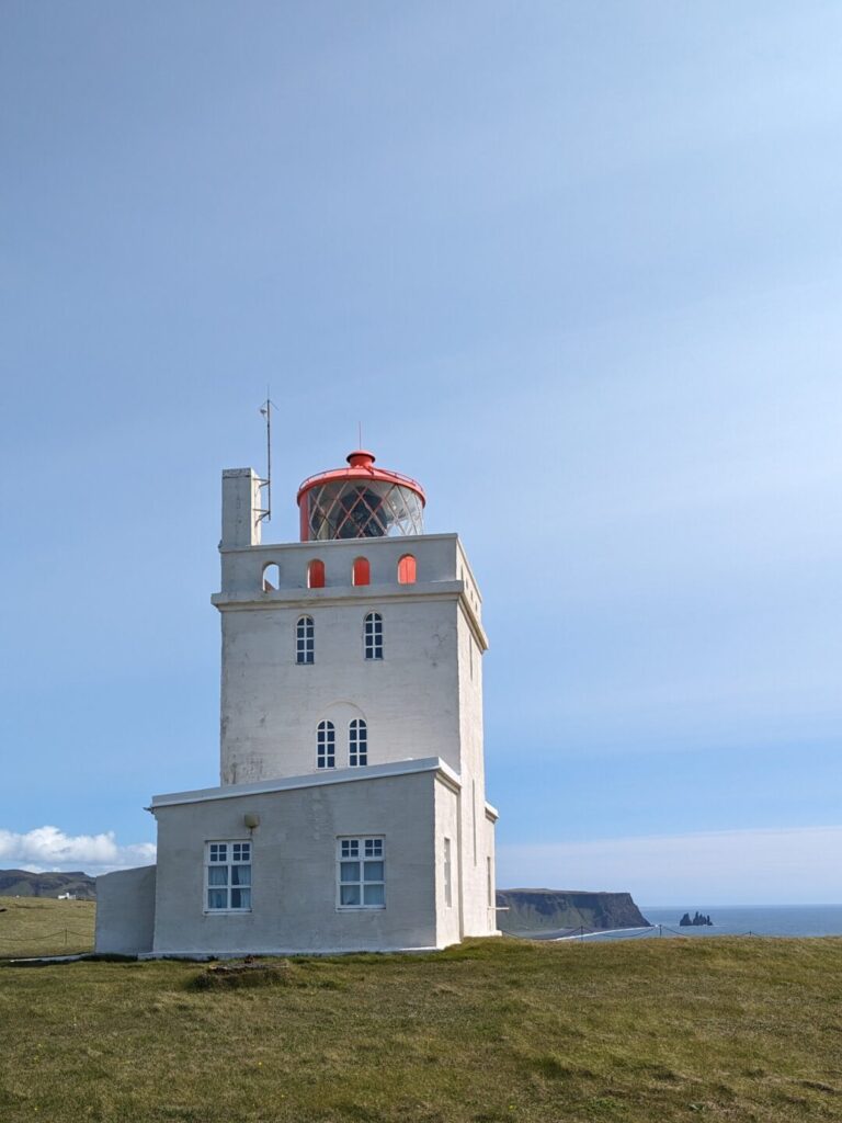 White lighthouse with an orange light on top sitting in a grassy field at the edge of a ocean front cliff