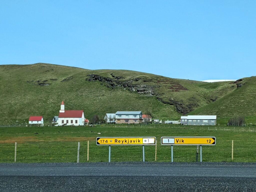 Grassy hills and field with a church and some small buildings in front of the hills. In the foreground there are two signs, one pointing left for Reykjavik and one pointing right for Vik