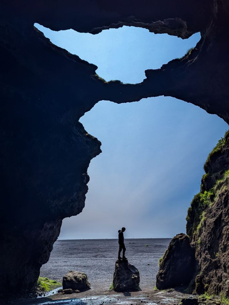 A cave that has holes shaped like yoda while looking out from the inside. a man stands on a rock in the opening looking down