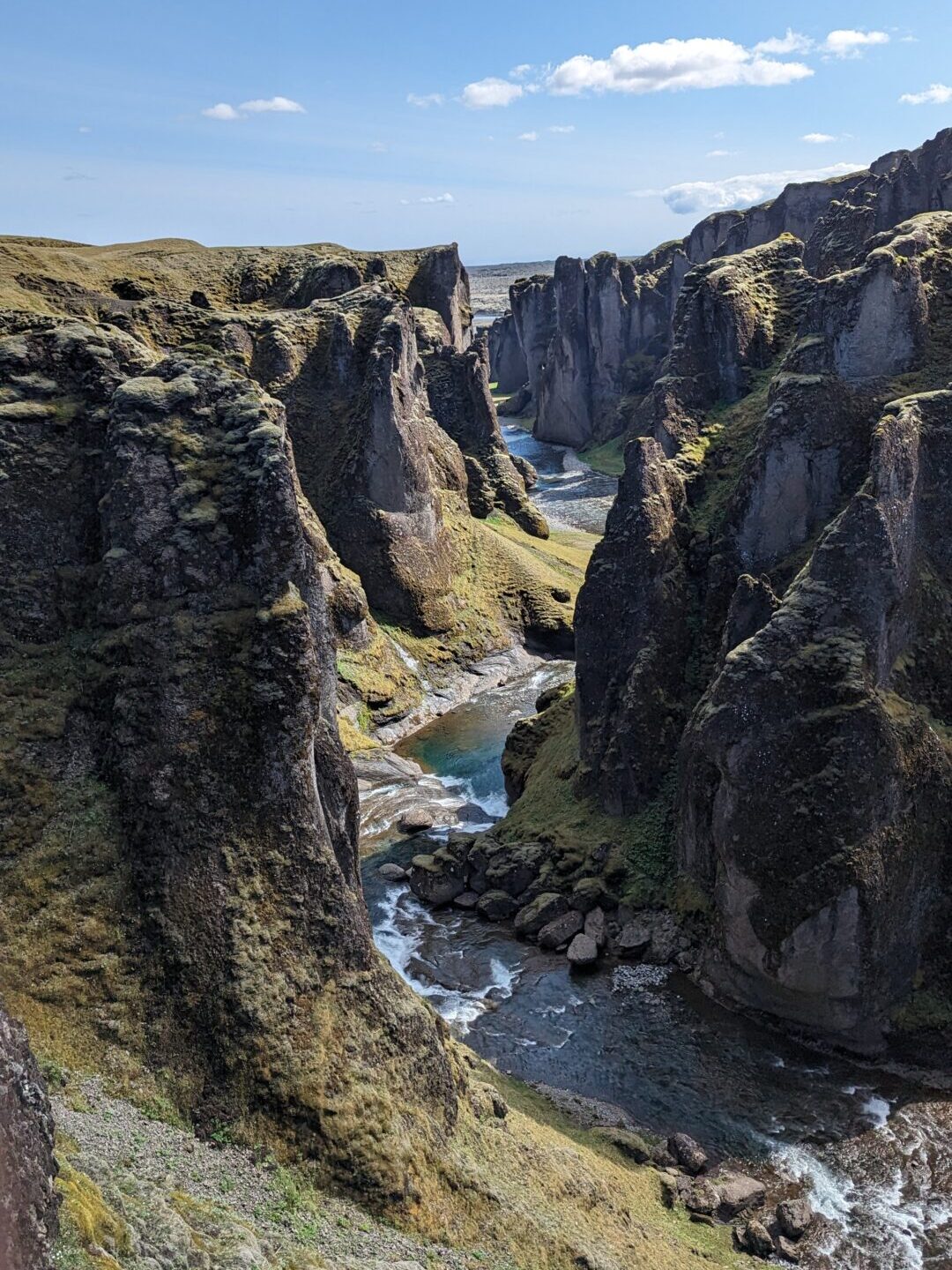 Mossy canyon with a river running down the center. The sky is bright blue with puffy clouds