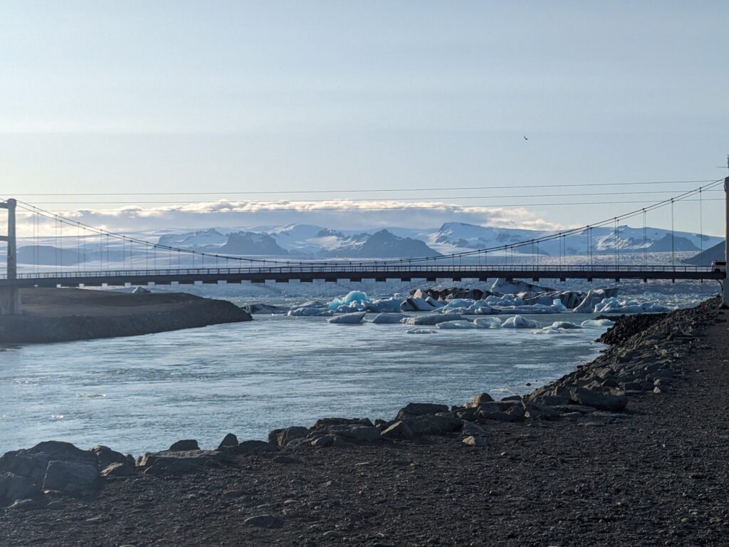 Icebergs floating on a glacial lake in the distance behind a bridge, with snow capped mountain in the background.