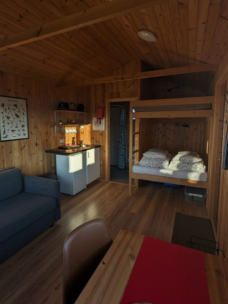 Inside of a wood panneled cabin. there is part of a couch visible on the left next to a counter and shelf with some coffee mugs. On the right side in the foreground is a table and chair and in the back there is a bunkbed with folded covers and a door to the bathroom to the left.